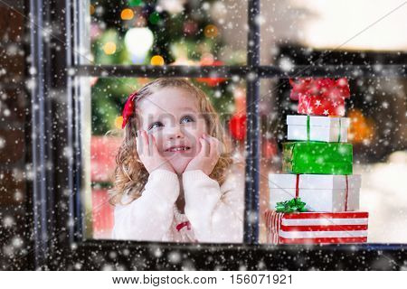 Little girl waiting for Santa at home window on Christmas eve. Kids opening Xmas presents. Child under Christmas tree with gift boxes. View from outside. Winter snowy day at home.