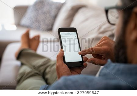 Closeup of a man hand holding cellphone with internet browser on screen. Man with spectacles relaxing sitting on couch. Closeup of mature latin man using smartphone to checking email at home.