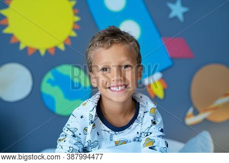 Portrait of smiling boy sitting in space decorated bedroom with the solar system and planets made of cardboard attached to the wall. Happy kid wearing an astronaut pajamas while looking at camera.