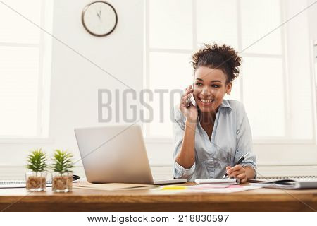 Happy smiling african-american business woman at work talking on phone and taking notes, sitting at her working place in office, copy space