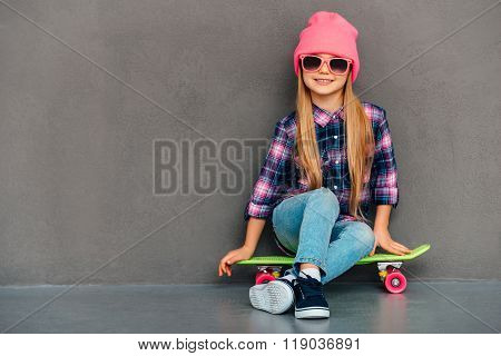 Carefree cutie with skateboard. Full length of cheerful little girl looking at camera with smile while sitting on skateboard against grey background