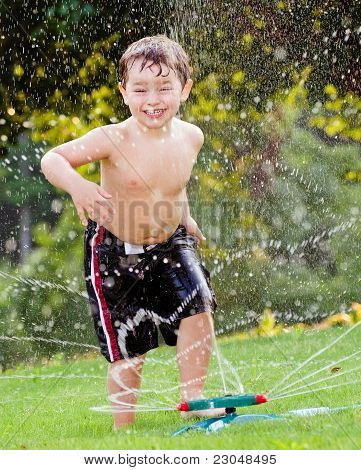 Young boy or kid cools off by playing in water sprinkler at home in his back yard on hot summer day