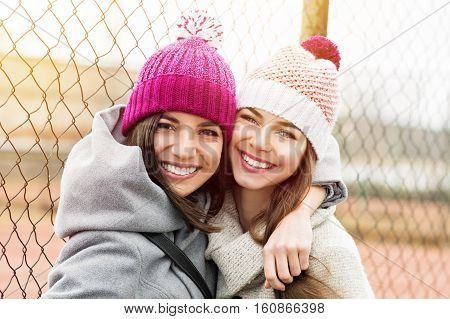 Two female besties in knit beanies and coats hugging and smiling. Two young best friends enjoying outdoors, loving and hugging each other. Vibrant color, natural light, medium retouch.