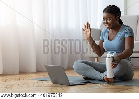 Smiling Black Woman In Sportswear Having Online Fitness Training With Laptop At Home, Waving Hand At