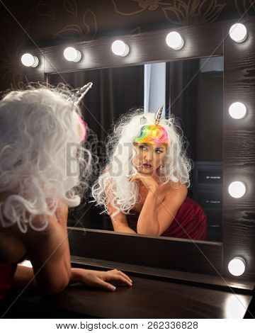 Freaky Young Woman In Unusual Wig Looks At Himself In The Mirror In Dressing Room. Portrait Of Stran