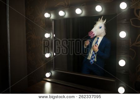 Glamour Guy In Elegant Suit And Comical Mask Looks At Himself In The Mirror In Dressing Room. Freaky