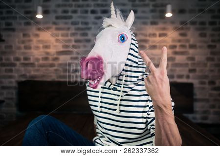 Freaky Young Man In Comical Mask Sits On Bed In Stylish Apartment. Funny Unicorn In Casual Clothes I