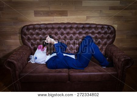 Funny Unicorn In Elegant Suit Lies On Sofa. Unusual Man Drinks Wine From The Bottle At Home. Freaky 