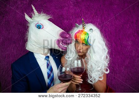 Portrait Of Freaky Couple On Purple Background. Strange Young Woman In Unusual Wig Drinking Wine Wit