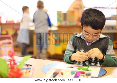 Boy In Glasses Moulds From Plasticine On Table In Kindergarten