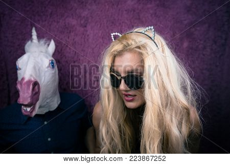 Freaky blonde girl in sunglass and kitty ears with funny guy in horse head mask on the purple background. Fantasy unicorn keep calm with girlfriend. Unusual young people sits together.