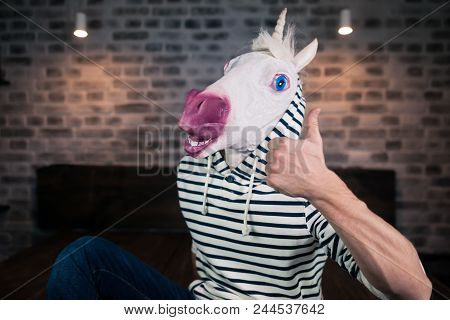 Freaky Young Man In Comical Mask Sits On Bed In Stylish Apartment. Funny Unicorn In Casual Clothes I