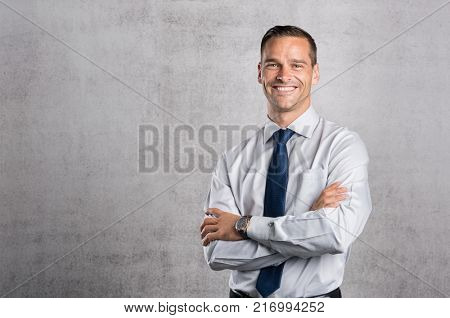 Happy businessman looking at camera on grey background with copy space. Handsome young business man standing against a grey wall with crossed arms. Successful formal man smiling with copy space.