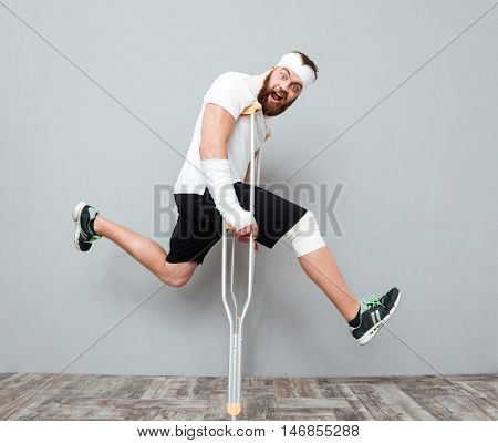 Freaky crazy young man jumping with crutches and shouting over gray background