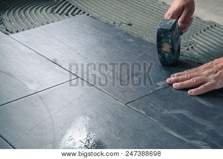 Worker placing ceramic floor tiles on adhesive surface, leveling with rubber hammer