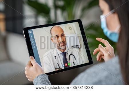 Sick woman wearing protective mask during video call with mature doctor. Back view of patient with surgical face mask talking during conference call with her physician, staying at home in quarantine. 