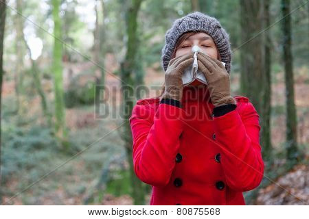 Young woman suffering from a cold or flu blowing her nose on a white paper handkerchief on a forest wearing a red overcoat, a beanie and gloves during winter 