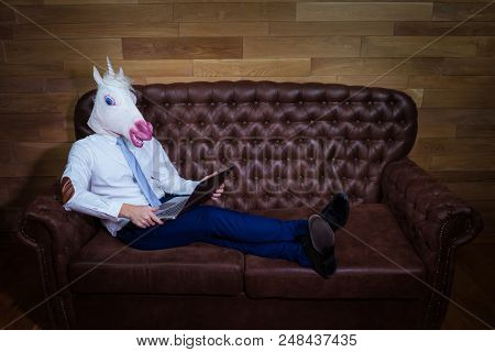 Unusual Man Working At Home Office. Freaky Young Manager In Comical Mask On Background Of Wooden Wal