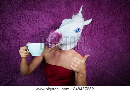 Funny Girl Unicorn Drinks Tea And Shows Thumbs Up Gesture. Freaky Young Woman In Comical Mask Stands