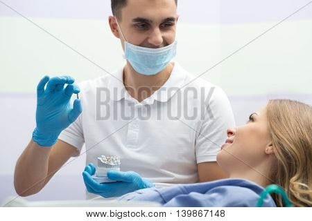 Young girl in blue shirt and dentist in a white uniform with blue latex gloves and a blue mask. He holds an implant in the right hand and teeth mould in the left hand. They look at each other. Horizontal.