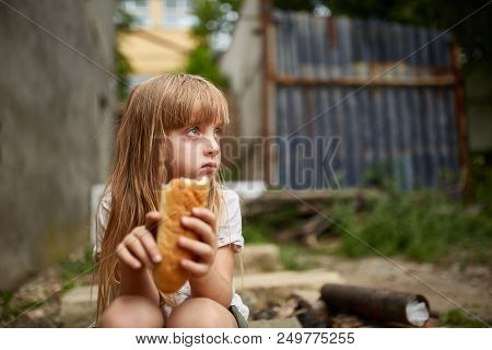 Portrait Of Poor Hungry Homeless Girl Eating A Piece Of Bread In The Dirty Alley, Selective Focus. P