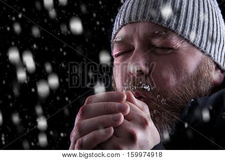 Freezing cold man standing in a snow storm blizzard trying to keep warm. Eyes closed and blowing warm air into his hands. Wearing a beanie hat and winter coat with frost and ice on his beard and eyebrows. Looking to the left.