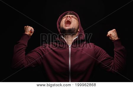 Furious, angry, freaky guy in a loose red sweater on a black background. A sick guy screaming and clenching fists in anger and self-loathing. Obsession, drug cravings, addiction concept.